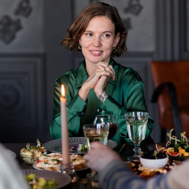 Mulher de camisa verde sorrindo à mesa de jantar com velas e convidados – a arte de receber com sofisticação
