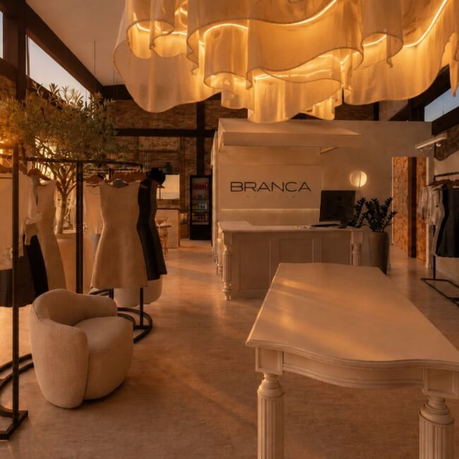 Interior loja Branca Boutique Itaipava Petrópolis luz dourada pôr do sol teto orgânico araras manequins mesa central ambiente